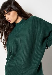 Maglione a coste di un verde scuro con collo alto, vestibilità oversize e dettagli con bottoni laterali ai polsini. Texture liscia e forma rilassata.