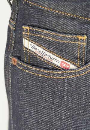 Dunkelblaue Jeans aus Denim mit sichtbaren hellbraunen Nähten und einem weißen Etikett mit der Aufschrift "Diesel Industry", das teilweise in eine Gesäßtasche gesteckt ist.
