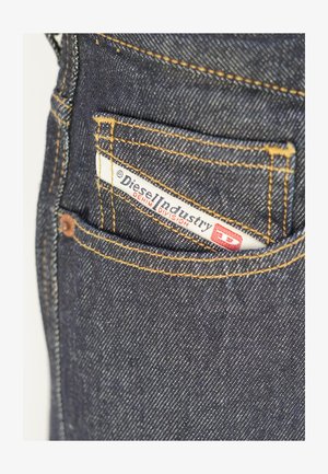 Jeans in denim blu scuro con cuciture visibili color marrone chiaro, e un'etichetta bianca con la scritta "Diesel Industry" parzialmente inserita in una tasca posteriore.