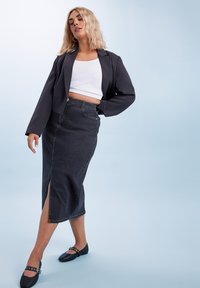 Blazer negro recortado sobre un top blanco corto, acompañado de una falda midi de mezclilla oscura con una abertura lateral y zapatos planos negros con hebillas.