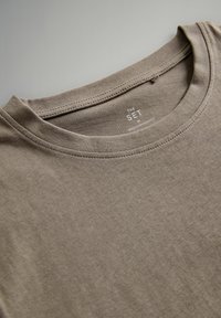 Ruda medvilnė marškinėliai su apvalia iškirpte, turintys paprastą tekstūrą ir matomas siūles. Etiketėje užrašyta "THE SET" ir "MADE IN BANGLADESH."