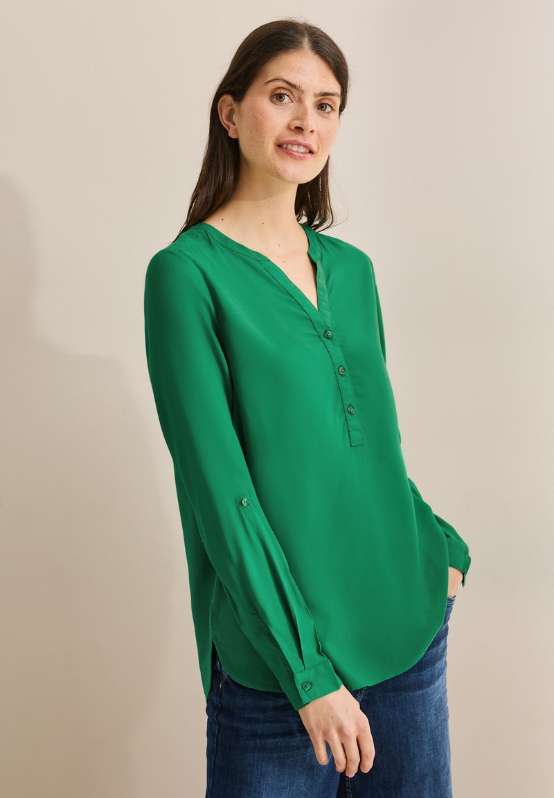 Blusa de poliéster verde con escote en V, mangas largas y puños abotonados. Presenta un dobladillo curvado y un brillo sutil. Se lleva con vaqueros azules.