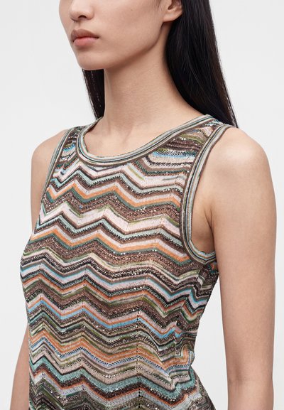 Femme aux longs cheveux foncés portant un haut sans manches à motif zigzag multicolore avec des accents métalliques.