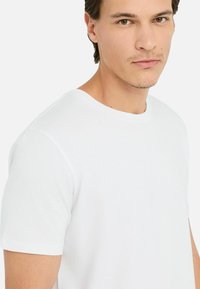 Junger Mann mit kurzem dunklem Haar, der ein schlichtes weißes T-Shirt mit Rundhalsausschnitt vor einem weißen Hintergrund trägt und leicht außerhalb der Kamera schaut.