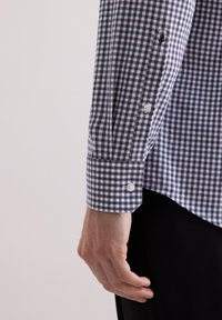 Camicia a quadri navy e bianca con polsini bottonati, caratterizzata da una trama di tessuto liscia e un orlo leggermente curvo. Mano visibile.
