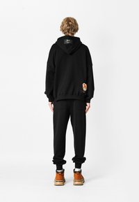 Sudadera negra con capucha grande, hombros caídos y parche de oso bordado en la parte posterior. Combinada con pantalones negros y zapatos marrones.