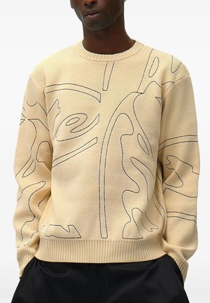 Uomo che indossa un maglione beige lavorato a maglia con motivi astratti di linee nere e pantaloni neri su sfondo bianco.