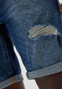 Džínové šortky v tmavě modré barvě, s poškozenou nášivkou a třepenými okraji. Ohrnuté manžety a decentní šicí detaily.