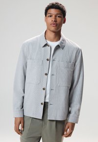 Young man wearing light grey textured button-up jacket over white shirt and olive green pants, standing against a plain background.