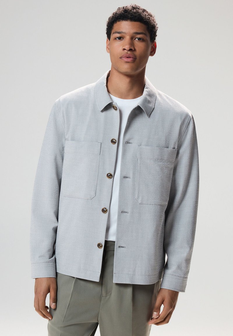 Young man wearing light grey textured button-up jacket over white shirt and olive green pants, standing against a plain background.