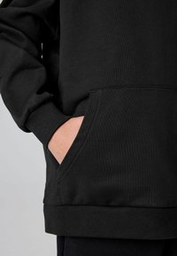 Oseba v črni kapuci z eno roko delno v sprednjem žepu proti nevtralni ozadju.