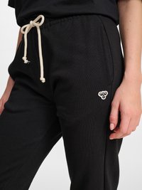 Schwarze Jogginghose mit weißem Kordelzug, der zu einer Schleife gebunden ist, Seitentasche und kleinem weiß besticktem Logo nahe der Hüfte. Hand ruht seitlich.