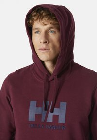 Bordový mikina s kapucí vyrobená z měkké látky, která má velké námořnické modré logo a detaily se šňůrkami na krku.