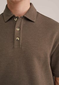 Polo de manga corta en una tela marrón texturizada con cuello y tapeta de tres botones, que presenta botones beige y un patrón de cuadros.