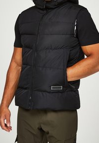 Černá prošívaná vesta na zip, s nastavitelnou kapucí a dvěma postranními kapsami. Obsahuje prošívanou strukturu a malou nášivku vpředu.