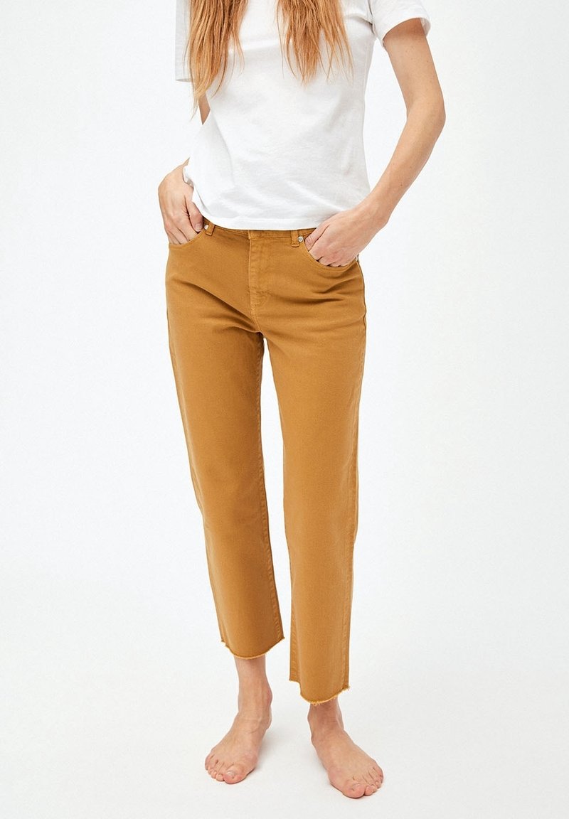 Femme portant un pantalon court moutarde et un t-shirt blanc, debout pieds nus avec les mains dans les poches sur un fond uni.