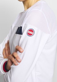 Biała bluza z siateczkową górną częścią, granatowymi akcentami oraz okrągłym czerwono-niebieskim logo "Colmar" na lewej rękawie.