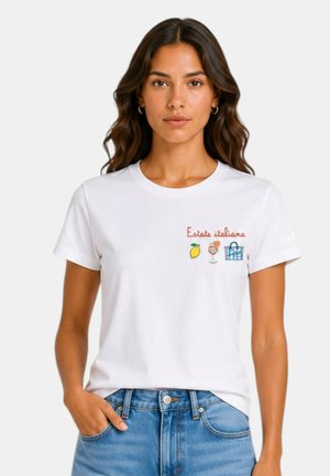 Donna con capelli lunghi e mossi che indossa una t-shirt bianca con testo "Estate italiana", limone, bevanda e ricamo di borsa, abbinata a jeans blu.