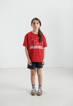 Rdeča bombažna majica z grafičnim dizajnom, ki vključuje oblike jajc in besedilo, kombinirana s temnimi denim kratkimi hlačami in čevlji z mešanimi vzorci.