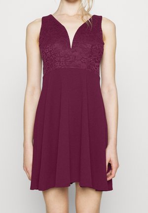 Vestido burdeos sin mangas con cuerpo floral de encaje y escote en V, llevado por una mujer sobre un fondo liso y claro.