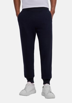 Mann trägt schwarze, gerippte Bündchenhose und weiße Sneakers, Hände in den Taschen, steht vor einem schlichten Hintergrund.