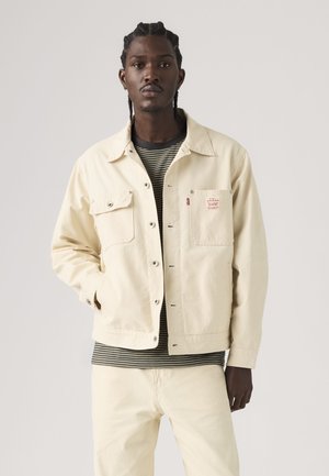 Young man with braided hair wears a cream jacket over a black and white striped shirt and cream pants against a plain background.