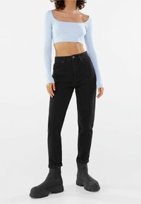 Kvinna i ljusblå långärmad croppad topp, höga svarta jeans som är uppvikta vid fållarna, och svarta kraftiga stövlar, som står mot en vit bakgrund.