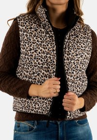 Gilet leopard stampa marrone e beige con colletto alto, zip frontale e maniche testurizzate marrone; indossato sopra una maglietta nera con bottoni.