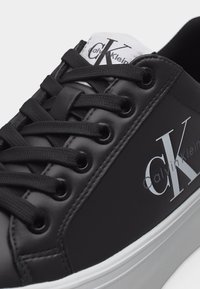 Μαύρο αθλητικό παπούτσι Calvin Klein με μαύρα κορδόνια, λευκό λογότυπο στο πλάι και λευκή ετικέτα στη γλώσσα με μαύρη σήμανση "CK".