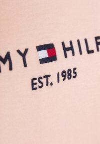 Lyserød stof har broderet teksten "TOMMY HILFIGER" i marineblå med et logo-symbolet i rød, hvid og blå samt etableringsåret 1985.