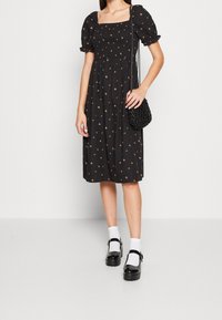 Robe noire midi avec manches bouffantes et décolleté carré, présentant un corsage smocké et de petites impressions florales. Portée avec des chaussures à plateforme noires.