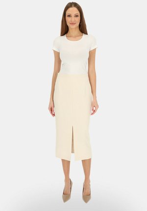 Kobieta stojąca, ubrana w biały top z krótkim rękawem, beżową spódnicę midi z rozcięciem z przodu i beżowe szpiczaste szpilki na jednolitym białym tle.