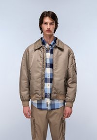 Beżowa kurtka bomberka z prążkowanymi mankietami i dołem, na niebieskiej koszuli w kratę zapinanej na guziki. Model ma na sobie jasnobeżowe spodnie cargo.