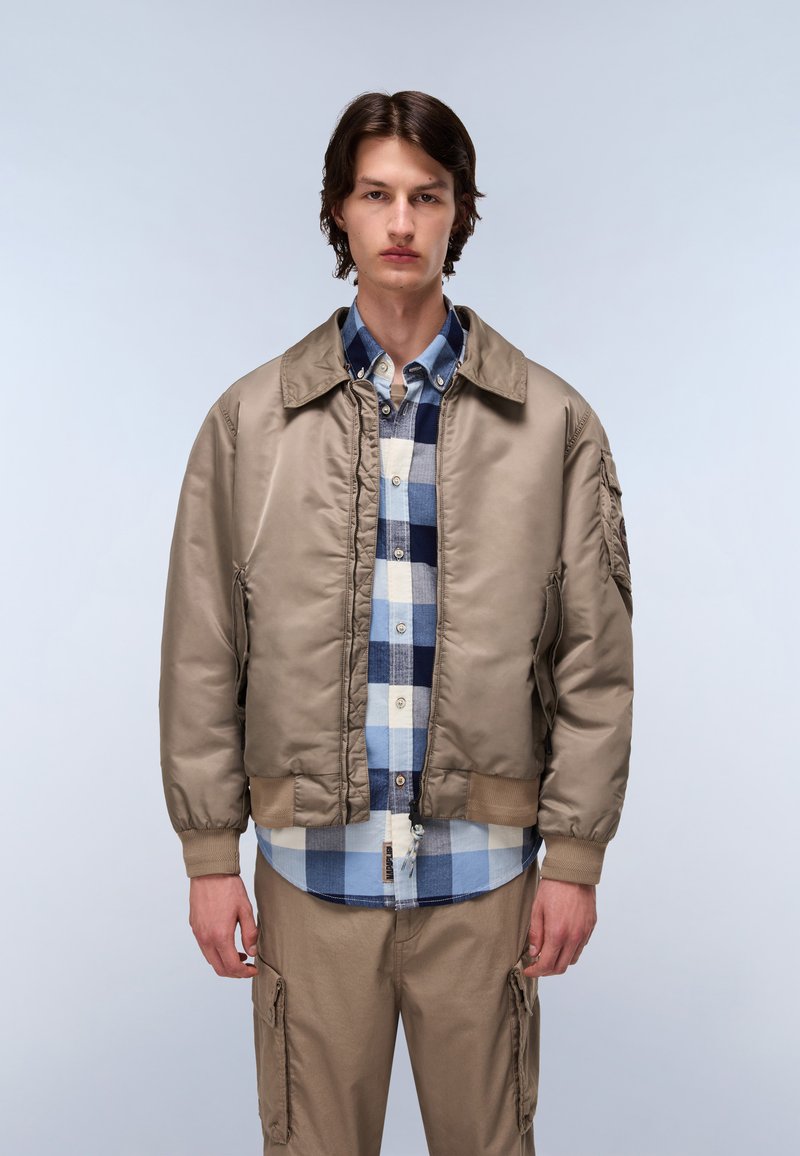 Beżowa kurtka bomberka z prążkowanymi mankietami i dołem, na niebieskiej koszuli w kratę zapinanej na guziki. Model ma na sobie jasnobeżowe spodnie cargo.