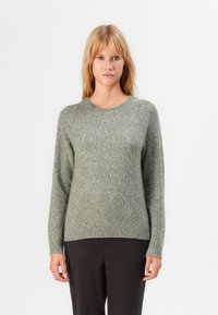 Woman with straight blonde hair wearing a grey knit sweater and black pants, standing against a white background facing forward.