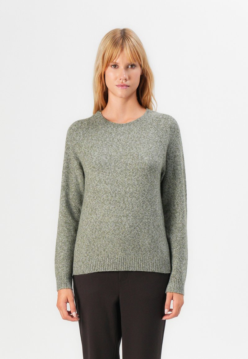 Femme aux cheveux blonds raides portant un pull en tricot gris et un pantalon noir, se tenant debout devant un fond blanc, faisant face.