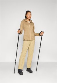 Turistický outfit, ktorý zahŕňa svetlo hnedú fleecovú bundu na zips s kapucňou a oranžovými akcentmi, béžové nohavice a čierne turistické topánky s trekingovými palicami.