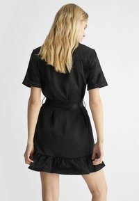 Abito corto nero con maniche corte, orlo arricciato e cintura in tessuto. Realizzato in materiale leggero, con una texture liscia. Mostrata vista posteriore.