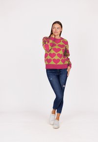 Vrouw in een groen en roze sweater met hartpatroon, donkere gescheurde jeans en witte sneakers, staand met haar hand op haar nek.
