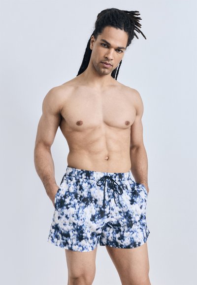 Shirtless man with dreadlocks wearing blue and white tie-dye swim shorts, standing with hands in pockets against plain background.