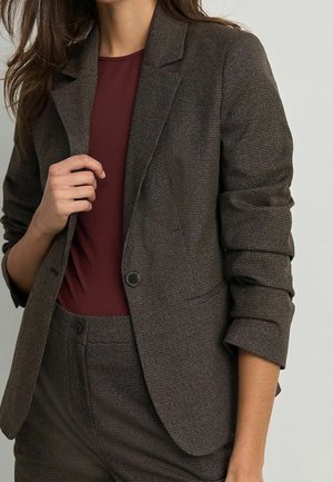 Femme portant un blazer à carreaux marron foncé et un pantalon assorti, associé à une chemise bordeaux, manches du blazer retroussées.
