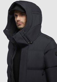 Giacca a vento navy con un ampio cappuccio, dotata di sezioni imbottite, chiusure con cordino e tasche laterali. Logo rotondo nero sul braccio sinistro.