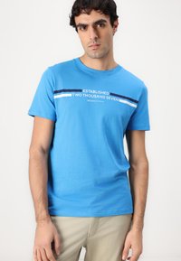 Mėlynas medvilnės T-shirtas su trumpomis rankovėmis, apvalia iškirpte ir grafiniu spaudiniu su užrašu "Įkurtas 2007" balta ir tamsiai mėlyna spalvomis.