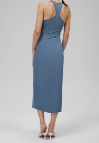 Niebieska bezrękawnikowa sukienka o dopasowanym kroju, długości midi, z bocznymi rozcięciami i gładką teksturą. Uzupełniona białymi sandałami na obcasie z odkrytymi palcami.