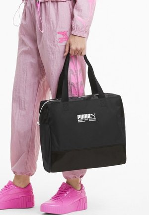 Osoba ubrana w różowy dres Puma i trampki trzyma czarną torbę typu tote Puma z białym logo za uchwyty.