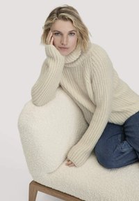 Cremfarbener, grob gestrickter Pullover mit hohem Kragen, geripptem Muster, kombiniert mit blauen Jeans, sitzend auf einem weichen, hellen Sofa.