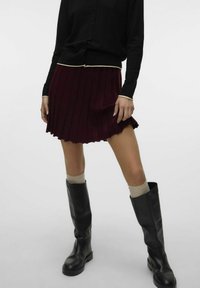 Jupe plissée bordeaux avec un ourlet en forme de vagues, associée à un cardigan noir avec des finitions crème et des bottes en cuir noir jusqu'aux genoux.