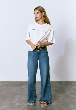 Camiseta de manga corta color crema con texto en verde y púrpura, combinada con pantalones de mezclilla azul de pierna ancha de cintura alta y un cinturón negro con tachuelas.