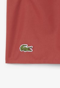 Tessuto rosso con logo di coccodrillo ricamato in verde e bianco. Superficie liscia con orlo pulito e dettagli di cucitura.