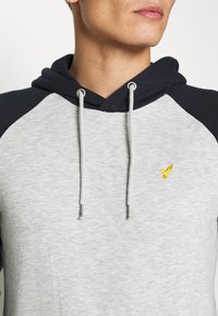 Helle graue Kapuzenjacke mit marineblauen Raglanärmeln. Sie hat eine strukturierte Oberfläche, silberne Ösen und ein kleines gelbes Vogel-Logo auf der Brust.